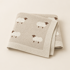 Beige knitted cotton baby blanket with cream sheep pattern, folded on a neutral background