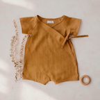 Golden tan linen baby romper flat lay with pampas grass and wooden ring toy